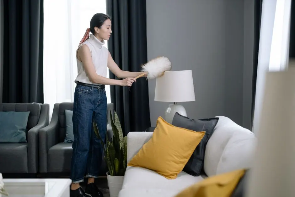Asian woman dusting a lampshade in a modern living room with yellow pillows.